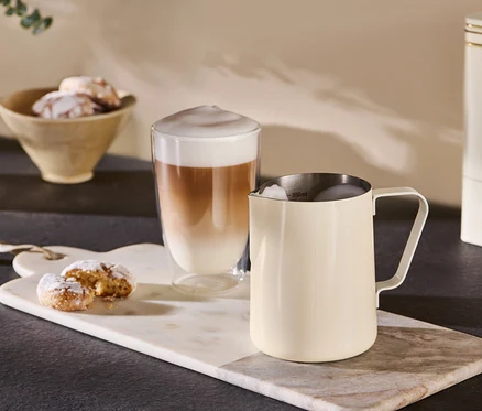 Ein beigefarbenes Milchkännchen steht neben einem Glas Kaffee auf einem Marmortablett.
