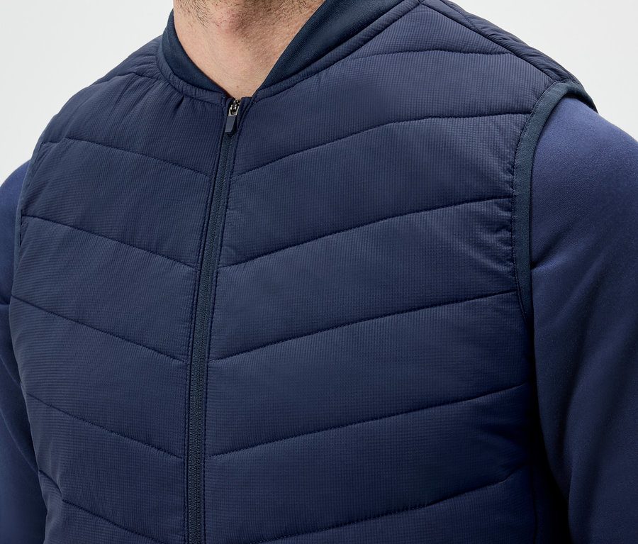 Ein Mann trägt eine dunkelblaue Steppweste mit Reißverschluss und ein Sweatshirt.