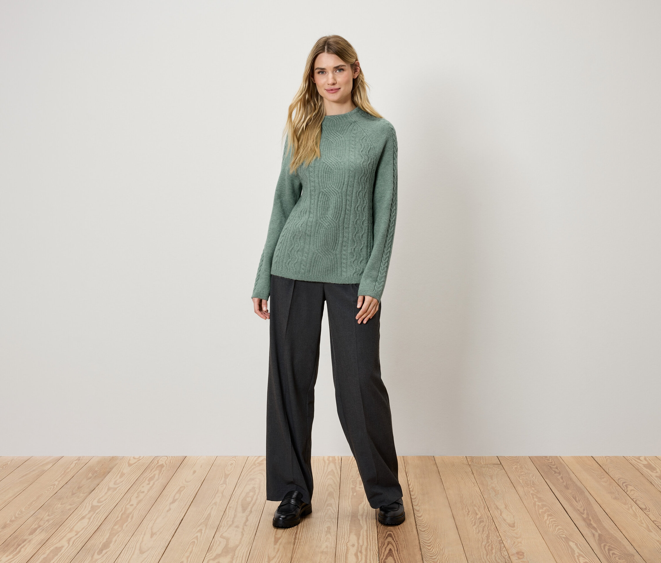 Eine blonde Frau steht auf einem Holzboden und trägt einen grünen Strickpullover und eine schwarze Hose.