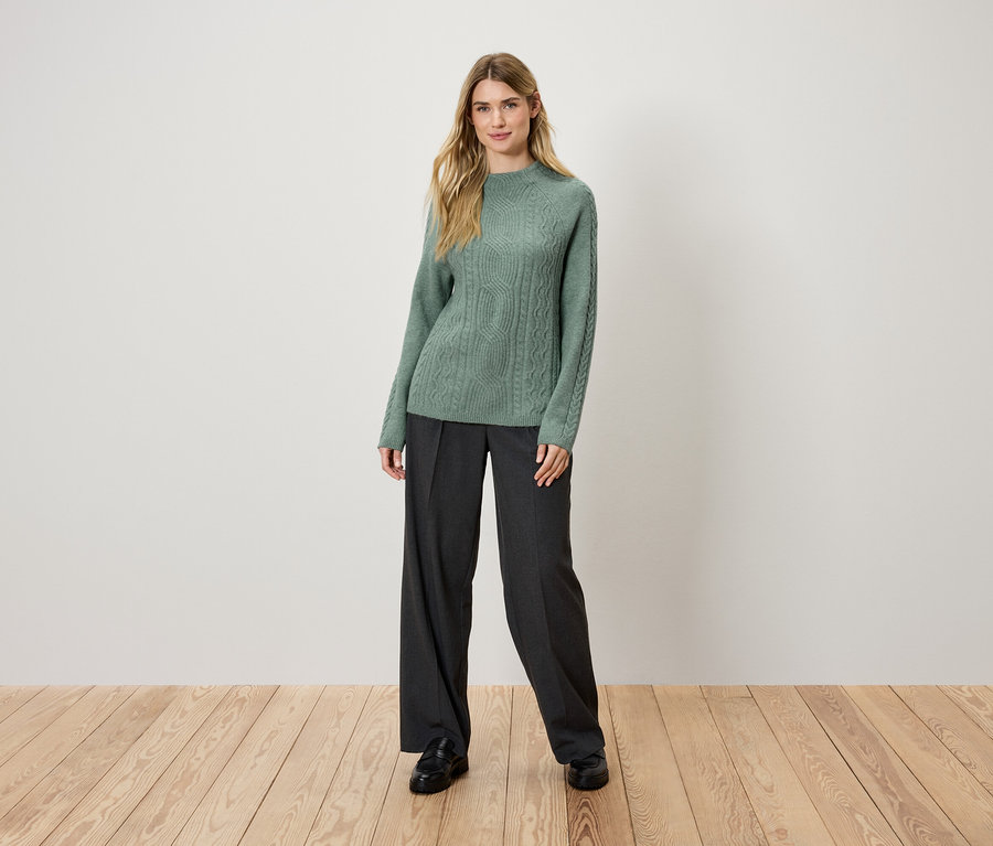 Eine blonde Frau steht auf einem Holzboden und trägt einen grünen Strickpullover und eine schwarze Hose.