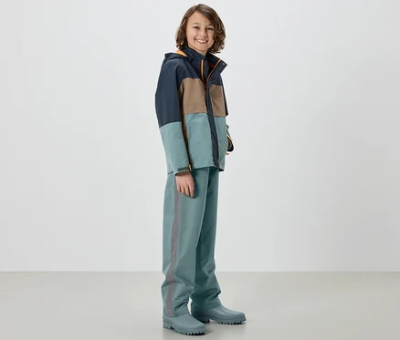 Ein Junge in einer Jacke und einer Kinder-Regenhose, Salbei, steht in Gummistiefeln.