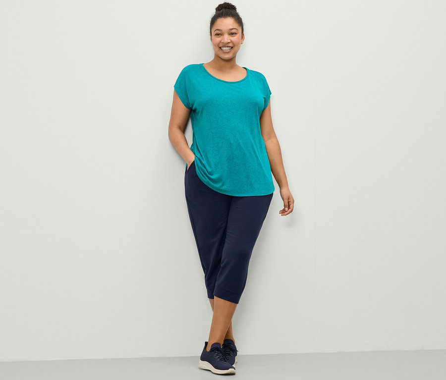 Frau posiert in türkisem Sportshirt und dunkelblauer 3/4-Sweathose.