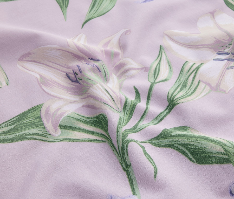 Nahaufnahme eines Stoffes mit blasslila Hintergrund, der mit weißen Lilien und grünen Blättern bedruckt ist.