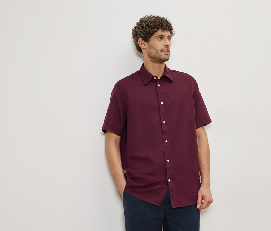 Mann mit lockigem Haar, der ein weinrotes, kurzärmeliges Hemd und eine marineblaue Hose vor einer weißen Wand trägt.