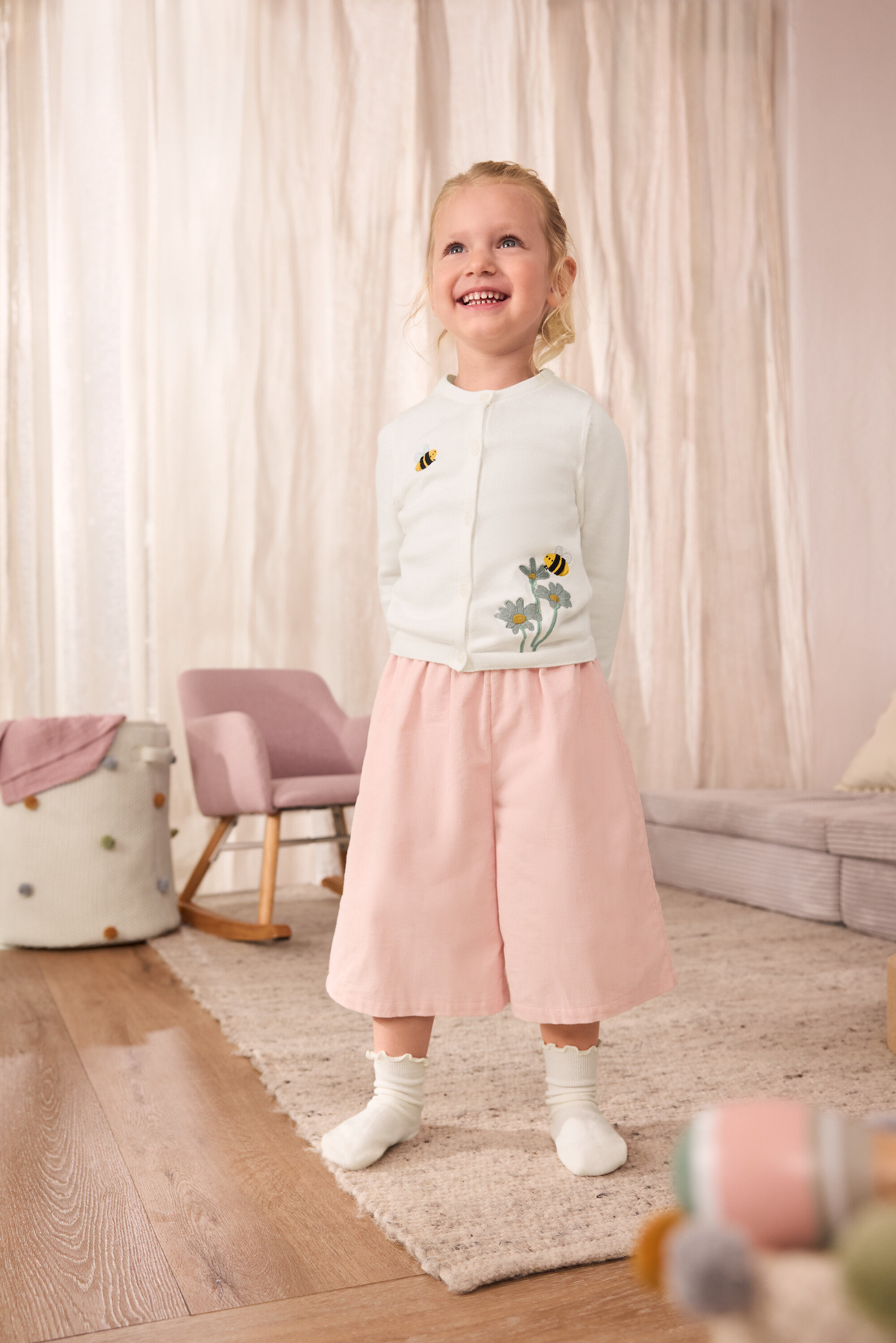 Ein lächelndes Mädchen trägt eine Kinder-Strickjacke mit aufgestickten Bienen, eine Kinder-Cord-Culotte und 5 Paar Kinder-Socken mit Rüschen.