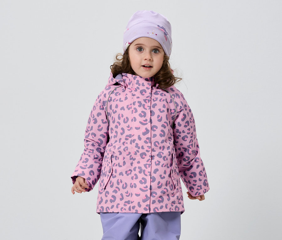 Ein kleines Mädchen mit lockigem Haar trägt eine rosa-lila Jacke mit Leopardenmuster, eine passende Mütze und lila Hosen vor einem weißen Hintergrund.