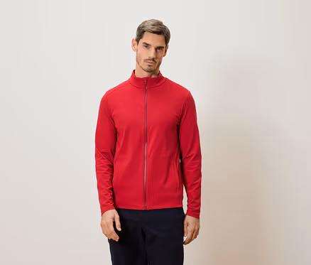 Mann steht vor einer Wand und trägt eine rote Jacke mit Reißverschluss.