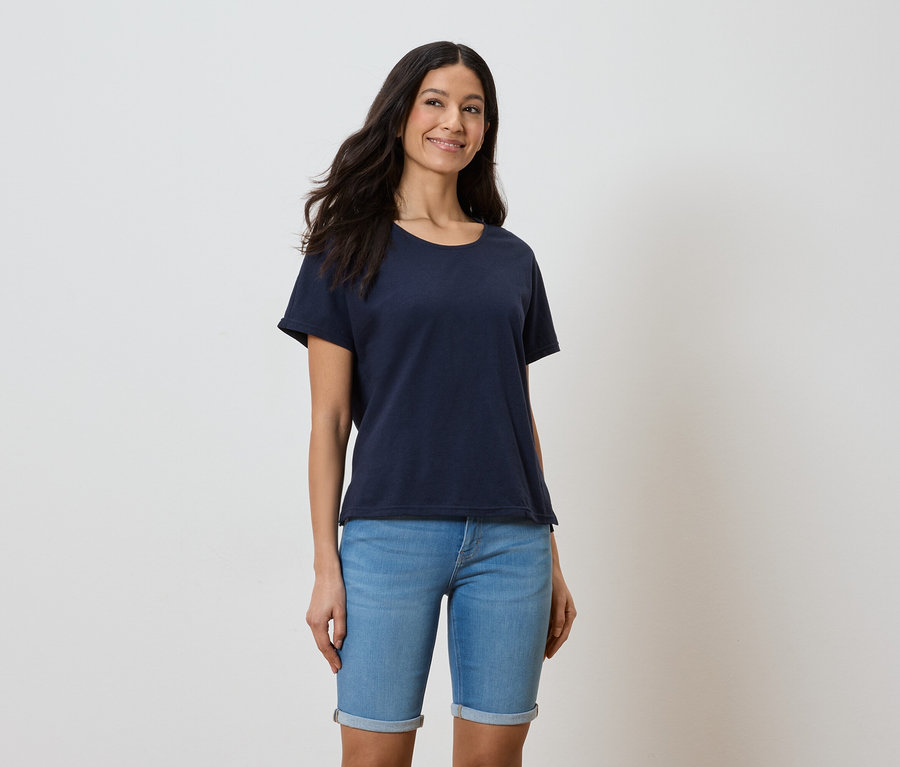 Frau mit dunklen, langen Haaren, die ein dunkelblaues T-Shirt und blaue Jeansshorts vor einem weißen Hintergrund trägt.
