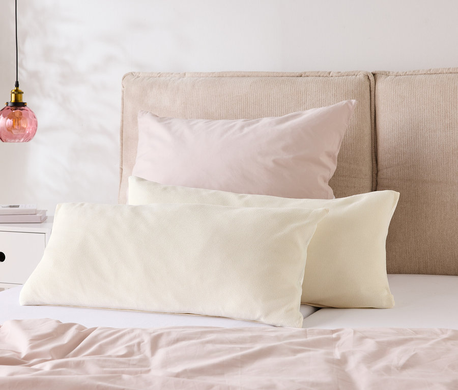 Ein Bett mit hellrosa Bettwäsche und zwei cremefarbenen Kissen liegt vor einem beigen Kopfteil. Auf dem Nachttisch steht eine Lampe.