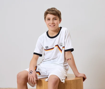 Lächelnder Junge mit Deutschland-Fußballtrikot und Shorts sitzt auf einem Hocker. Auf seinen Armen und Beinen sind Fußballtattoos.