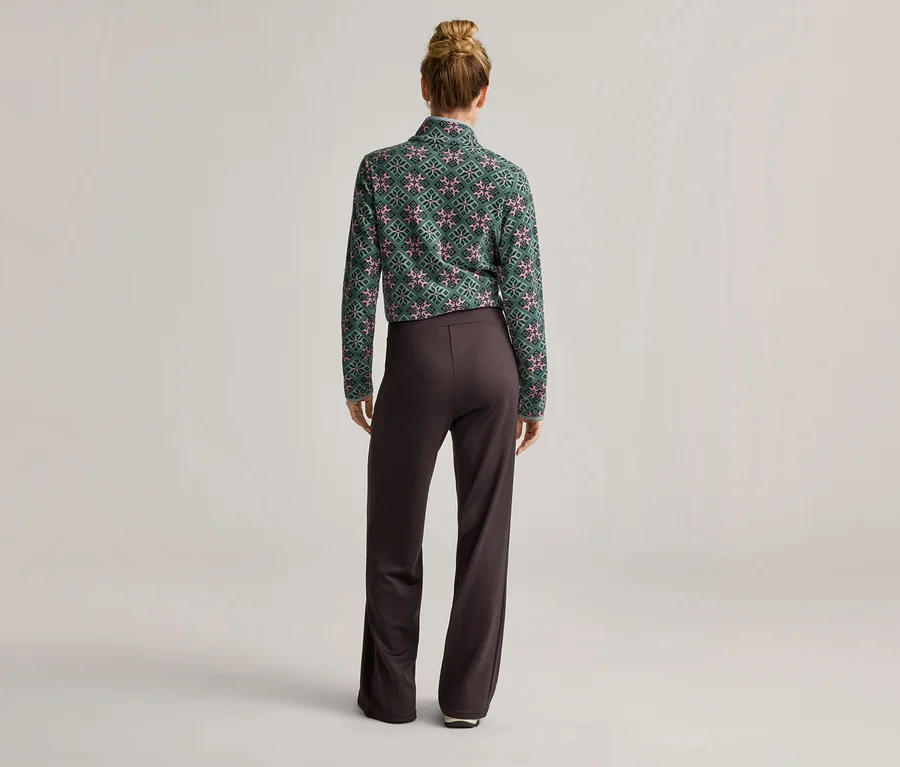Rückansicht einer Frau mit Duttfrisur, die ein grün gemustertes Langarmshirt und eine braune Hose trägt.
