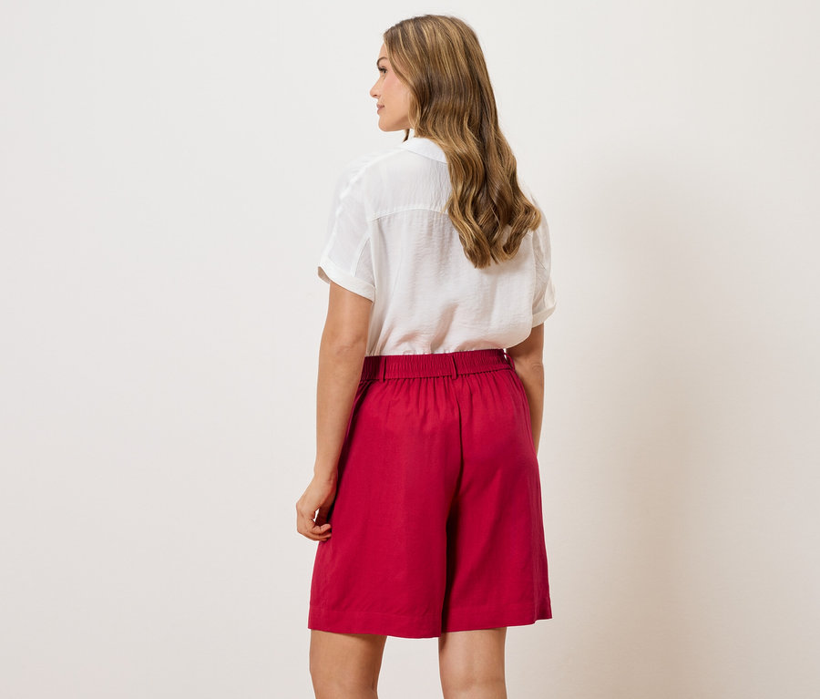 Rückansicht einer Frau mit gewelltem Haar, die eine weiße Bluse und rote Shorts trägt.