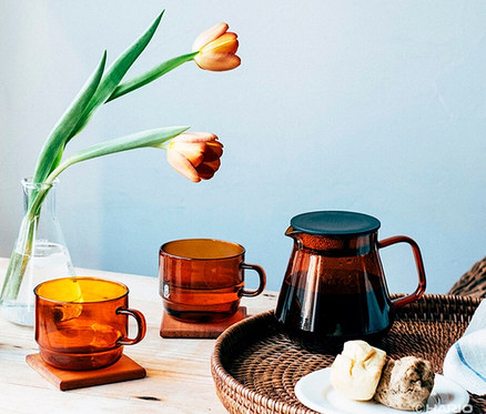 Auf einem Tisch steht ein HARIO »Colors« Tee- und Kaffee-Set in Amber, bestehend aus Kanne und Tassen, neben einer Vase mit Tulpen.