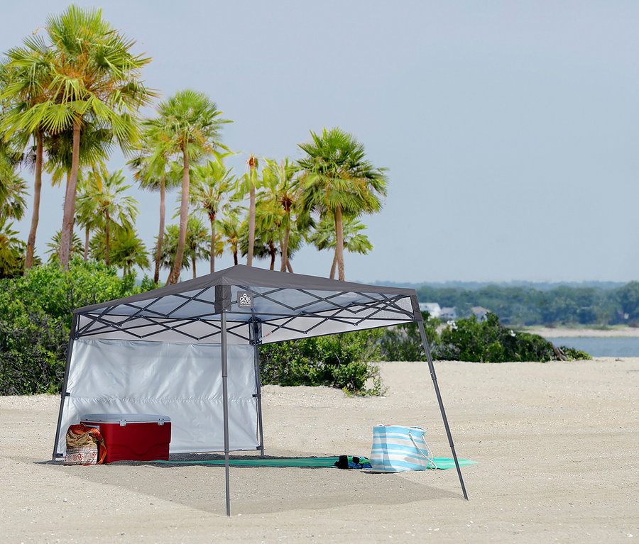 Am Strand steht ein graues Faltzelt mit Kühlbox, Tasche und Strandtasche davor. Im Hintergrund sind Palmen und das Meer zu sehen.