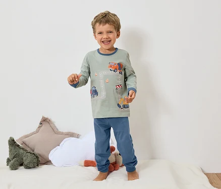 Ein lächelnder Junge steht barfuß auf einer weißen Oberfläche und trägt einen grauen Pyjama mit blauen Bündchen und einem Bagger- und Betonmischer-Motiv.