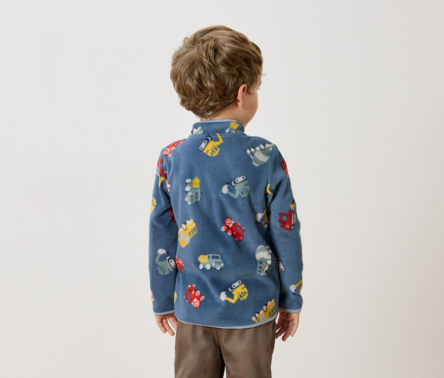 Rückansicht eines kleinen Jungen mit lockigem Haar, der einen blauen Fleecepullover mit bunten Fahrzeugmotiven und braune Hosen trägt.