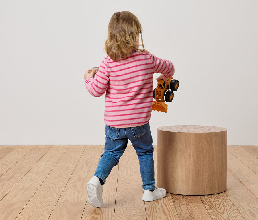 Ein Kind mit blonden Haaren steht mit dem Rücken zum Betrachter und hält einen Spielzeugbagger in der Hand.