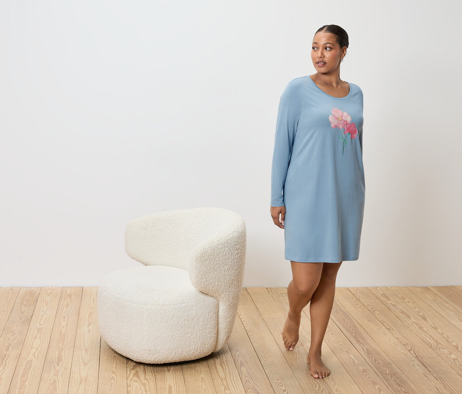 Frau im blauen Nachthemd mit Blumenmuster, neben einem weißen Sessel auf Holzboden.