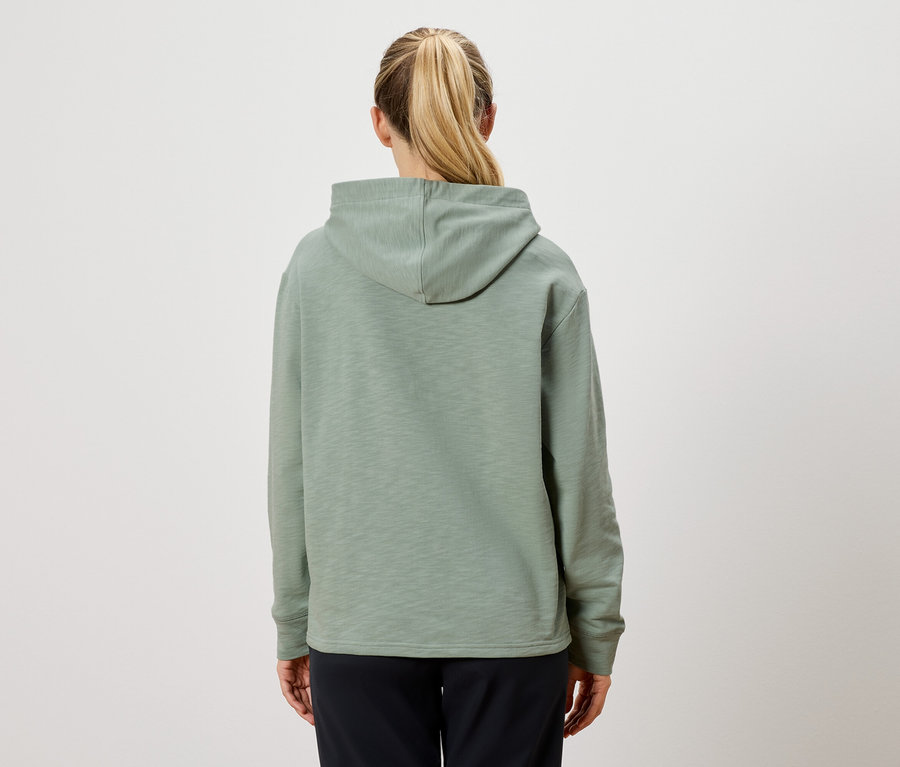Rückansicht einer Frau mit blondem Pferdeschwanz, die einen hellgrünen Kapuzenpullover und schwarze Hosen trägt.