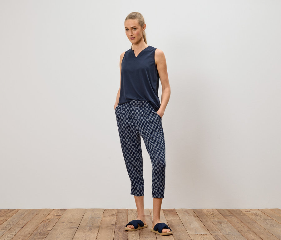 Frau steht mit Händen in den Hosentaschen, trägt ein marineblaues Top, gemusterte Hose und blaue Sandalen.