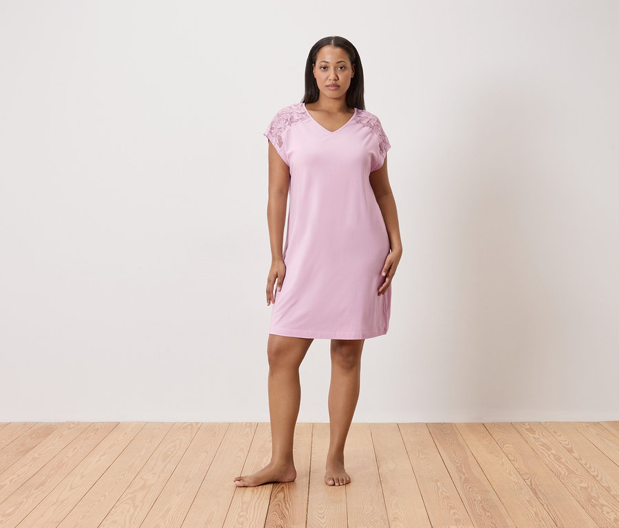 Frau steht barfuß auf einem Holzboden und trägt ein rosa Nachthemd mit Spitzenärmeln vor einer weißen Wand.