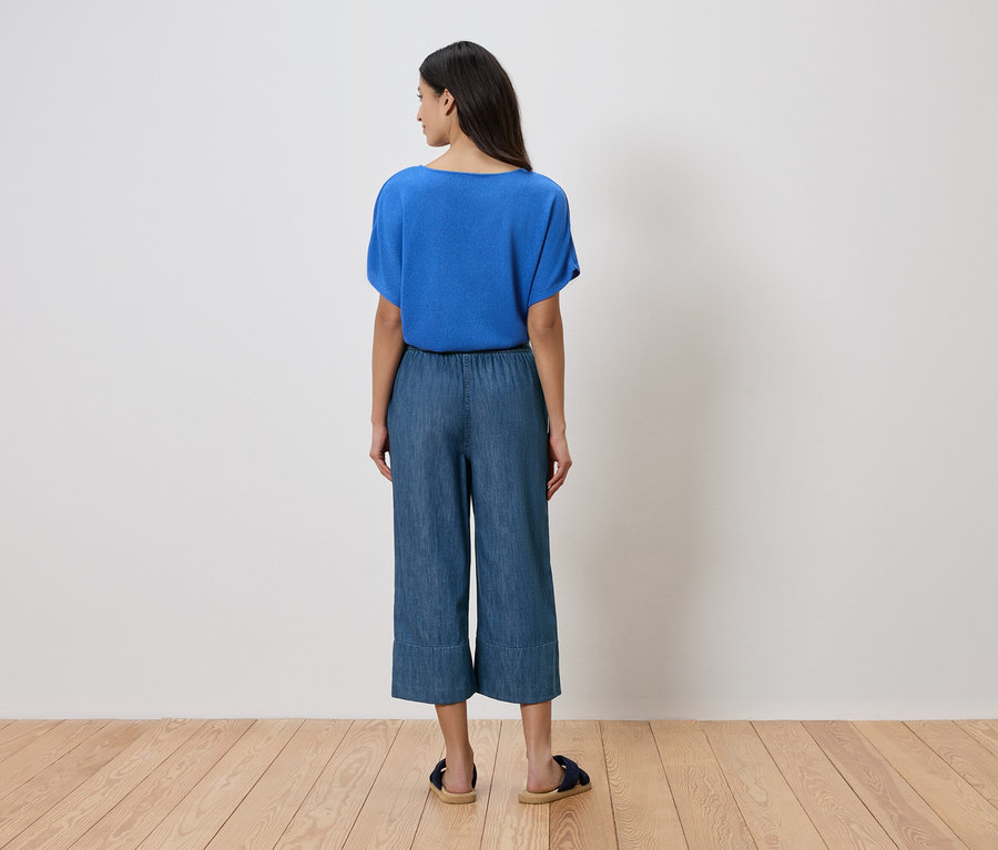 Frau von hinten in blauer Bluse, Culotte Jeans und Sandalen vor einer weißen Wand.