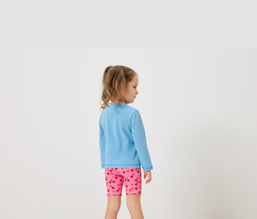 Mädchen von hinten in hellblauem Langarmshirt und pinken Shorts mit Schuppenmuster.