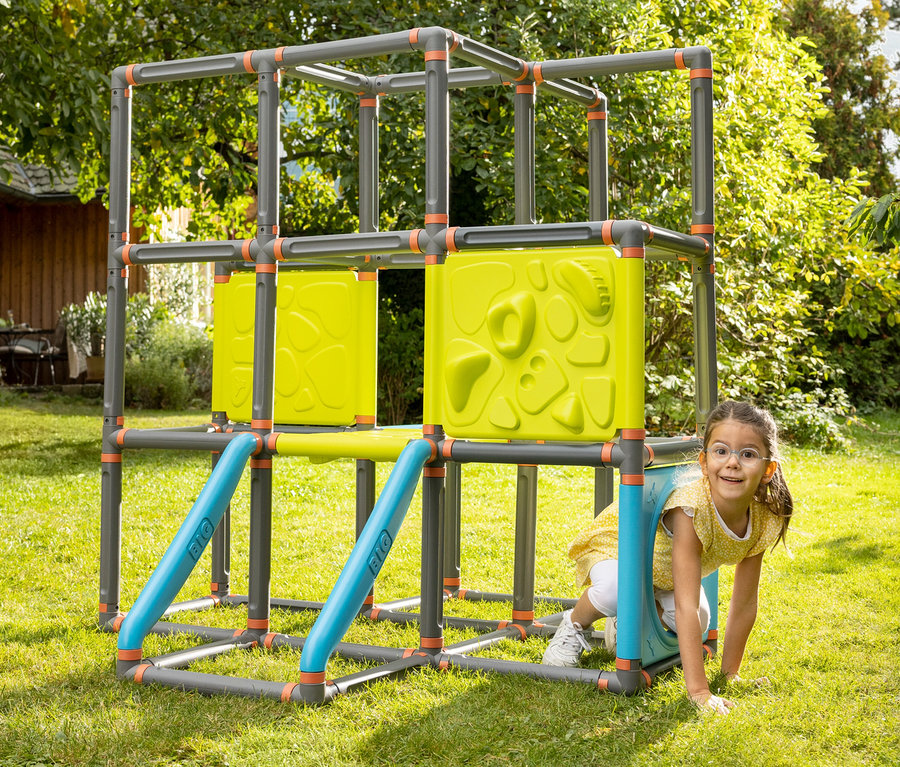 Mädchen kriecht durch ein graues Klettergerüst mit gelben Paneelen und blauen Stangen auf einer grünen Wiese.