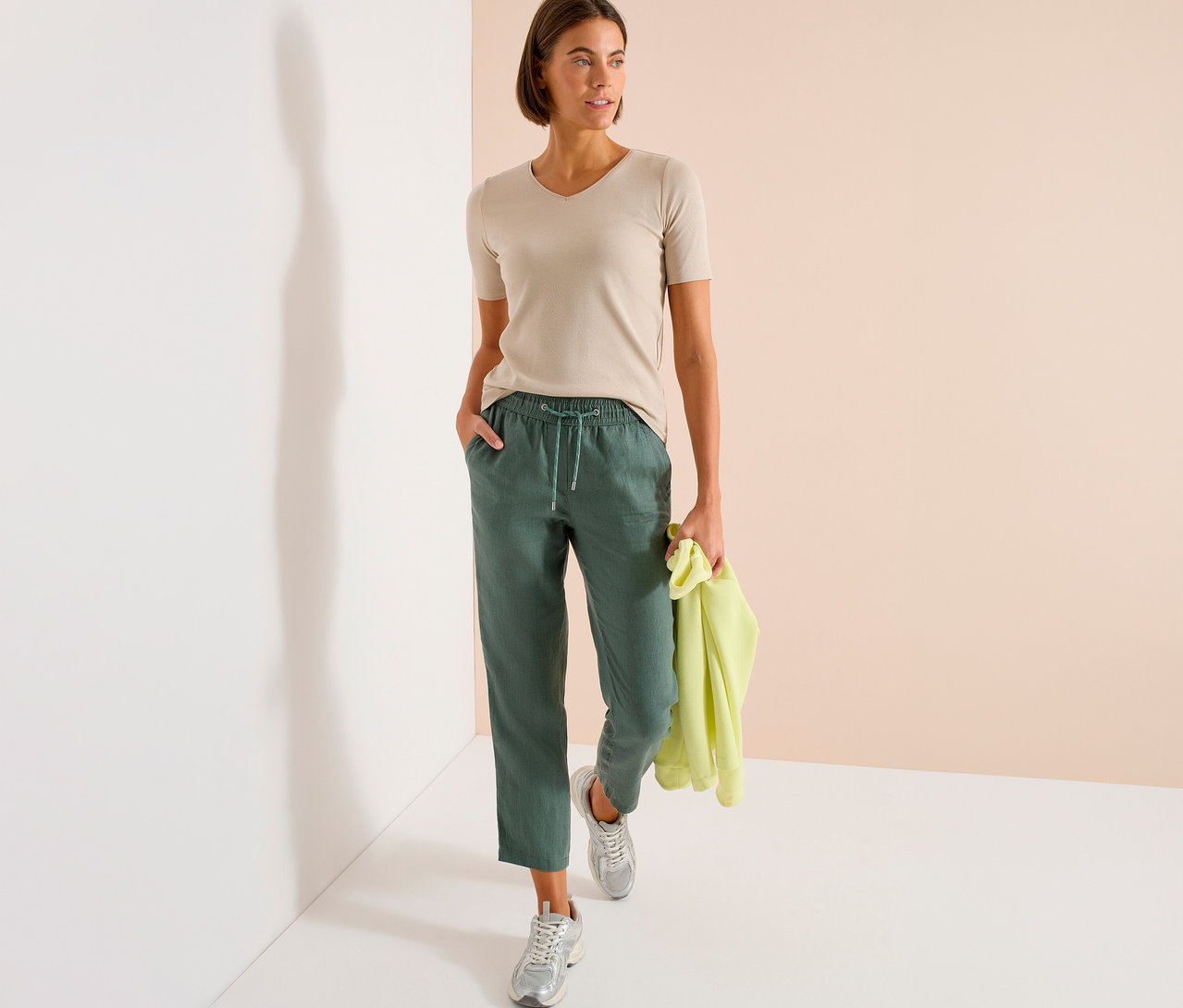 Frau in beigem T-Shirt und grüner Hose mit gelbem Pullover in der Hand, stehend.