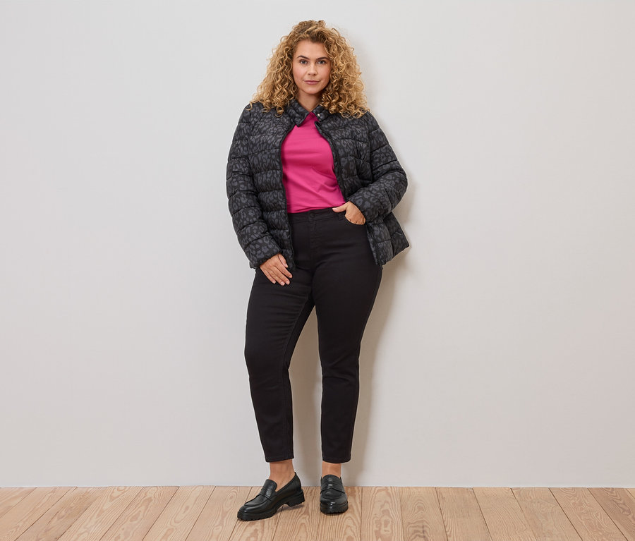 Frau mit lockigem Haar, trägt eine grau-schwarze, gesteppte Jacke über einem pinken Oberteil, kombiniert mit schwarzen Jeans und Loafern.