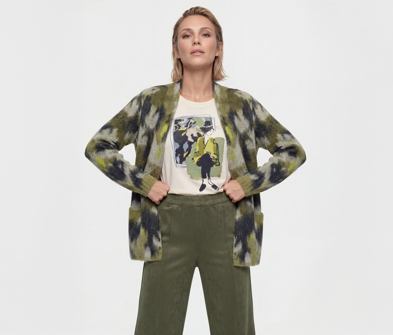 Eine Frau mit kurzen blonden Haaren steht mit den Händen in den Hüften und trägt einen offenen, grün-schwarz-gelb gemusterten Cardigan über einem cremefarbenen T-Shirt mit Grafik und olivgrünen weiten Hosen.