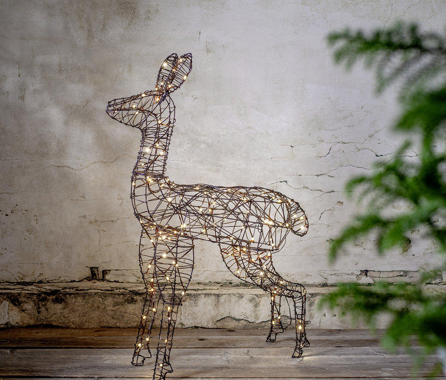 Eine Hirsch-förmige Drahtskulptur, beleuchtet mit warmweißen Lichtern, steht auf einem Holzboden vor einer strukturierten Wand. Rechts ist eine unscharfe grüne Pflanze.