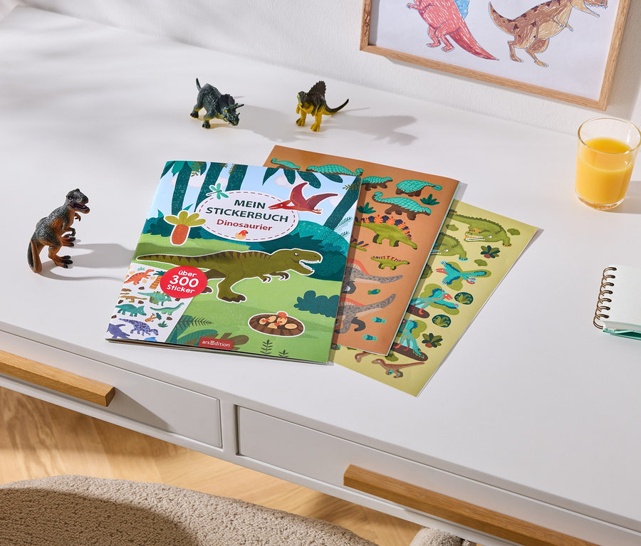 Ein weißer Schreibtisch mit einem Dinosaurier-Stickerbuch, das über 300 Sticker enthält, und zwei Stickerbögen. Daneben stehen drei kleine Dinosaurierfiguren, ein Glas Orangensaft, ein Notizbuch und zwei Buntstifte. Im Hintergrund hängt ein gerahmtes Dinosaurierbild an der Wand.
