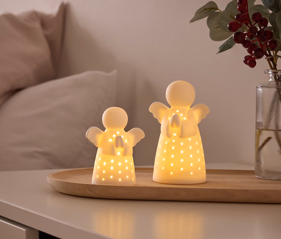 Zwei leuchtende, weiße Engelsfiguren mit ausgestanzten Löchern stehen auf einem Holztablett auf einem Nachttisch. Im Hintergrund sind ein Bett und eine Vase mit roten Beeren zu sehen.