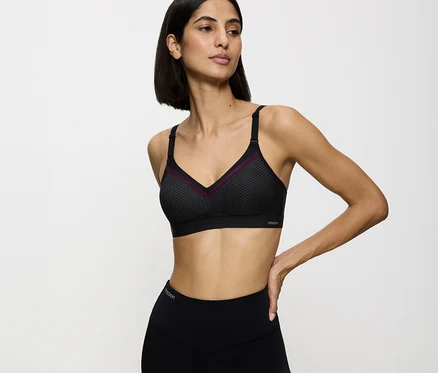 Frau trägt einen schwarzen TRIUMPH Sport-BH Triaction Free Motion N EX mit lila Zierleiste und schwarze Leggings, die linke Hand an der Hüfte.