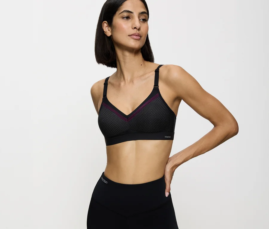 Frau trägt einen schwarzen TRIUMPH Sport-BH Triaction Free Motion N EX mit lila Zierleiste und schwarze Leggings, die linke Hand an der Hüfte.