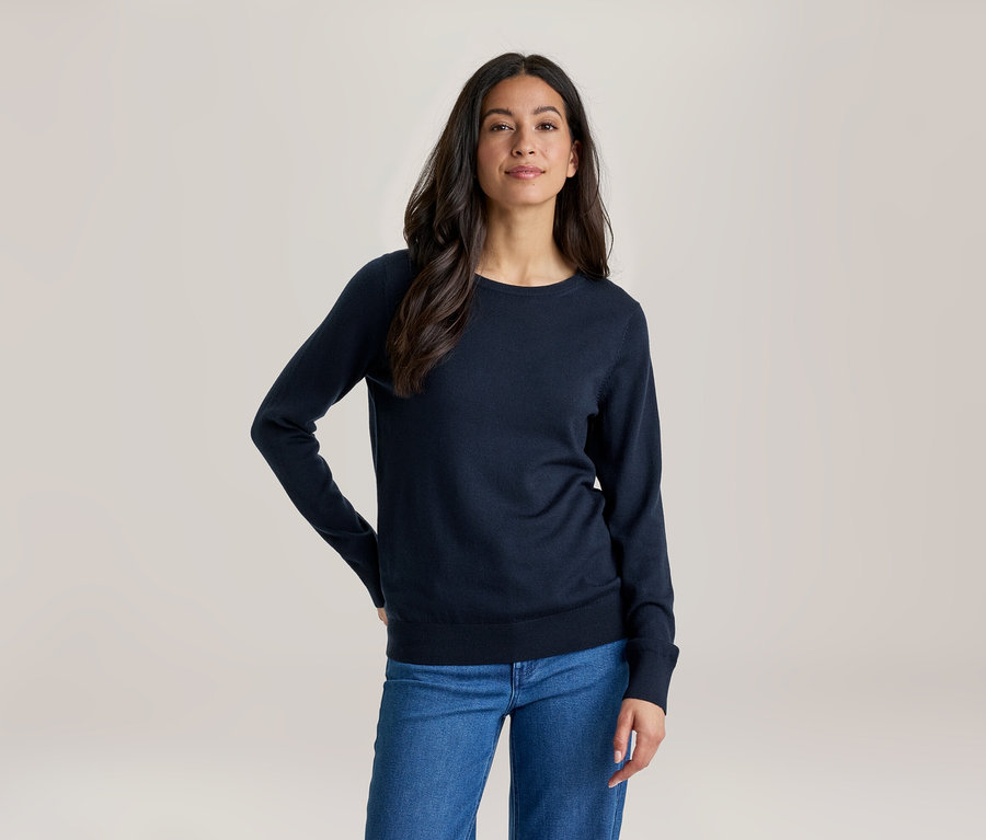 Eine Frau mit langen dunklen Haaren, die ein dunkelblaues Langarmshirt und blaue Jeans trägt, steht mit einer Hand in der Hüfte und blickt nach vorne.