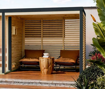 Ein offener Holzpavillon mit zwei Sesseln mit braunen Polstern und einem Baumstamm-Tisch, umgeben von üppigem Grün.