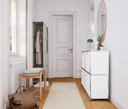 Heller Flur mit Holzboden und beigem Läufer. Links steht eine Massivholzbank mit Polster und Stauraum unter einem Fenster, daneben eine braune Reisetasche und ein offener Kleiderschrank. Rechts befindet sich ein weißes Sideboard mit einem runden Spiegel darüber.