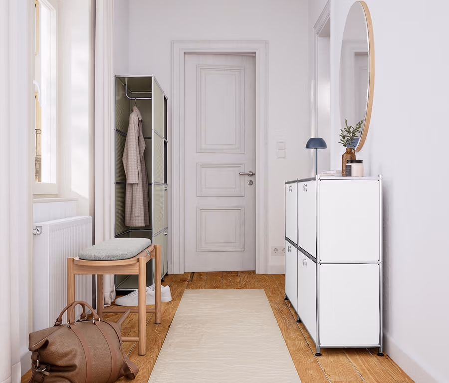 Heller Flur mit Holzboden und beigem Läufer. Links steht eine Massivholzbank mit Polster und Stauraum unter einem Fenster, daneben eine braune Reisetasche und ein offener Kleiderschrank. Rechts befindet sich ein weißes Sideboard mit einem runden Spiegel darüber.