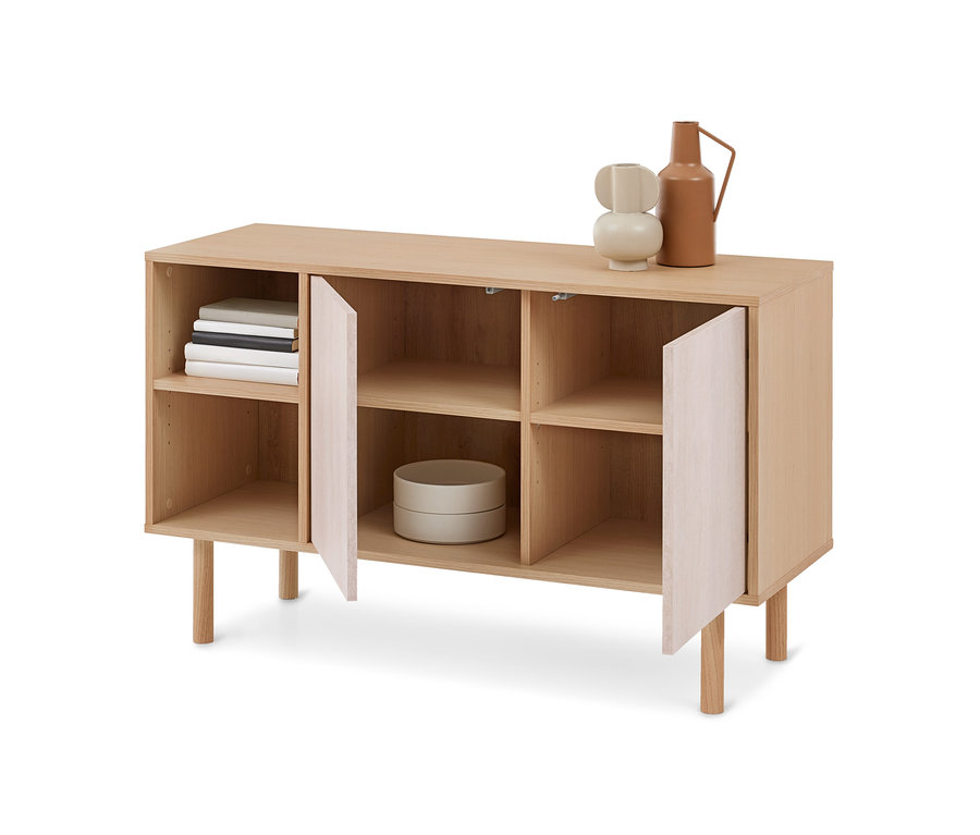 Helles Holz-Sideboard mit offenen Fächern und zwei leicht geöffneten Türen. Auf einem oberen Fach liegen Bücher, in einem unteren Fach steht ein hellgrauer Behälter. Obenauf stehen eine beigefarbene Vase und ein terrakottafarbener Krug.