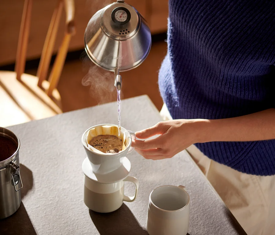 Frau gießt Wasser aus einem Kessel in einen weißen Kaffee Handfilter, Größe 101, mit Kaffeefilterpapier Gr. 101.