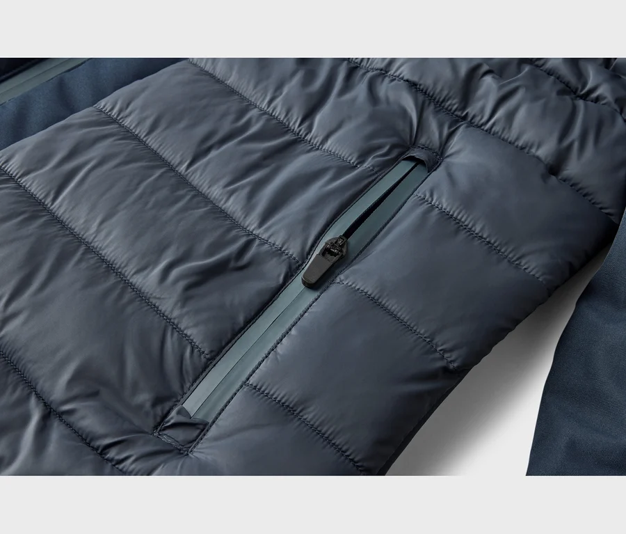 Nahaufnahme einer dunkelblauen Steppjacke mit horizontalen Nähten und einer diagonalen Reißverschlusstasche. Der Reißverschluss hat einen schwarzen Zipper und ein helleres blaues Reißverschlussband.