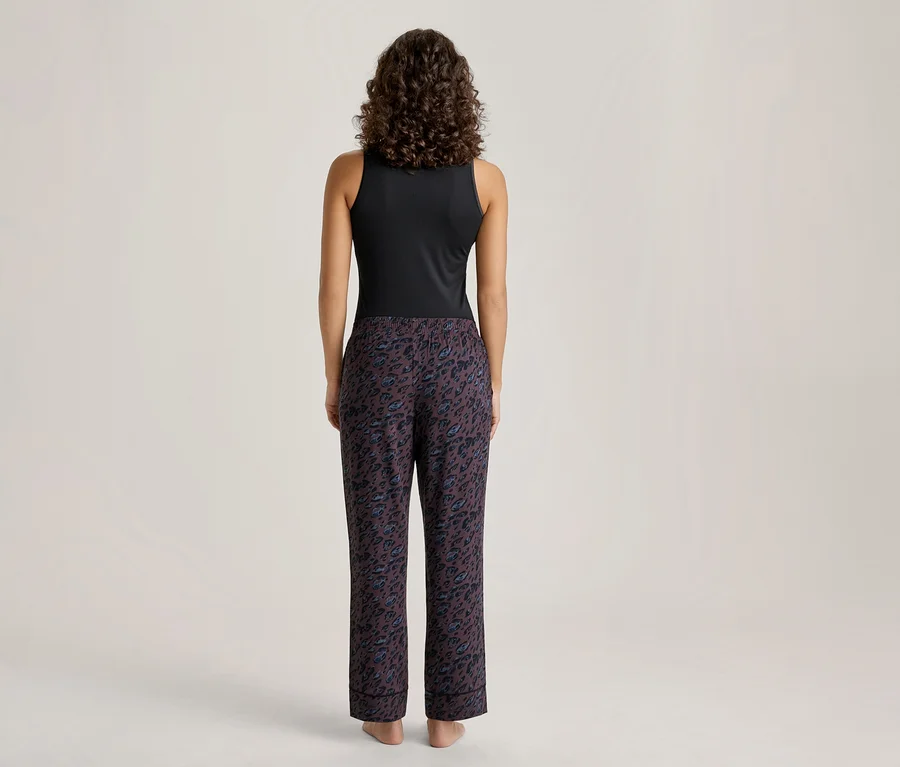 Eine Person mit lockigem Haar steht mit dem Rücken zum Betrachter und trägt ein schwarzes ärmelloses Oberteil sowie eine dunkle Hose mit Animal-Print. Die nackten Füße sind sichtbar.