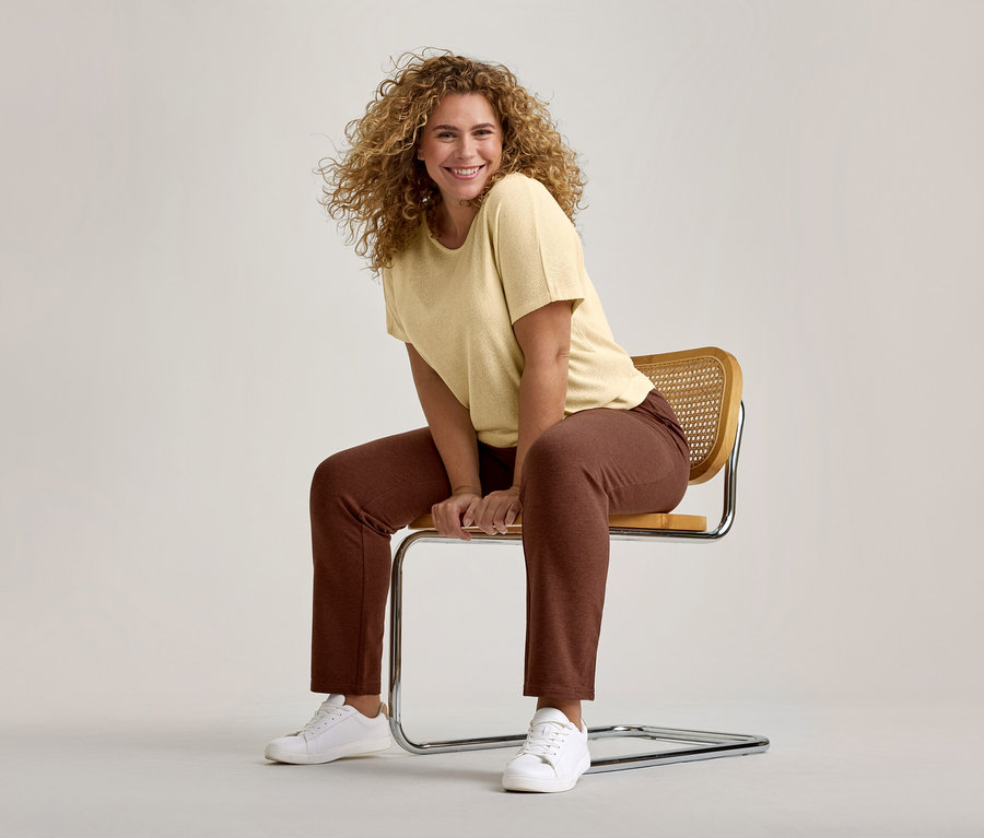 Lächelnde Frau mit lockigem Haar, die ein hellgelbes T-Shirt, braune Hose und weiße Sneaker trägt und auf einem modernen Stuhl sitzt.