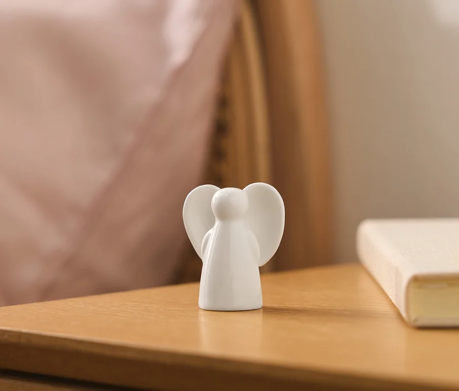 Eine weiße Keramik-Engelsfigur, von hinten gesehen, steht auf einem Holznachttisch, mit einem unscharfen rosa Kissen und einem hellen Buch im Hintergrund.