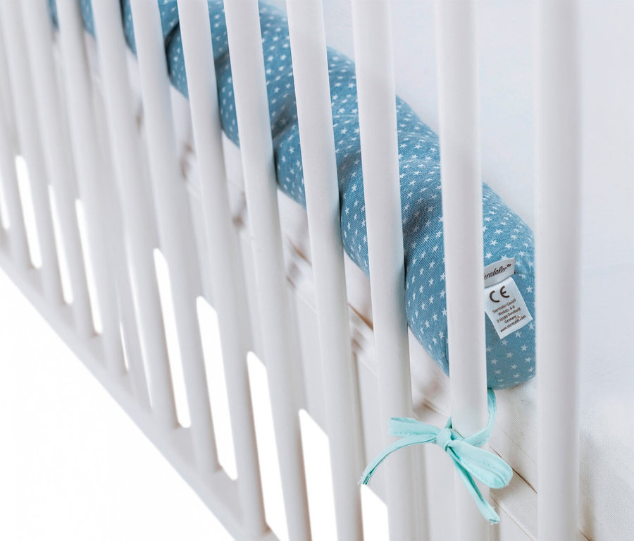 Blauer Bettschutz mit weißen Sternen an den weißen Gitterstäben eines Kinderbetts mit einer hellblauen Schleife befestigt.