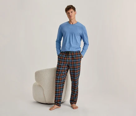 Mann steht, trägt ein blaues Langarmshirt und karierte Pyjamahose in Rot, Grün und Blau. Hände in den Hosentaschen, barfuß. Hinter ihm steht ein heller, texturierter Sessel.