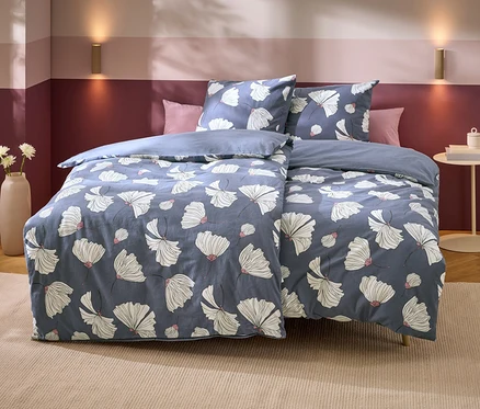Ein Bett mit blauer Bettwäsche mit weißen Blumenmustern und rosa Akzenten. Dahinter zwei rosa Kissen. Die Wand ist zweifarbig lila, mit zwei goldenen Wandleuchten. Links steht eine Vase mit weißen Blumen, rechts ein Beistelltisch mit Büchern.