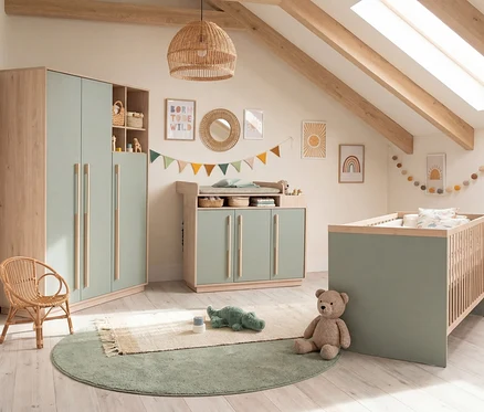 Gemütliches Kinderzimmer mit Möbeln in Salbeigrün und hellem Holz. Ein Kleiderschrank, Wickeltisch und Babybett sind zu sehen. Dekorationen umfassen Wandbilder, Girlanden und einen Rattansessel. Ein Teddybär und ein Krokodilspielzeug liegen auf dem Boden.